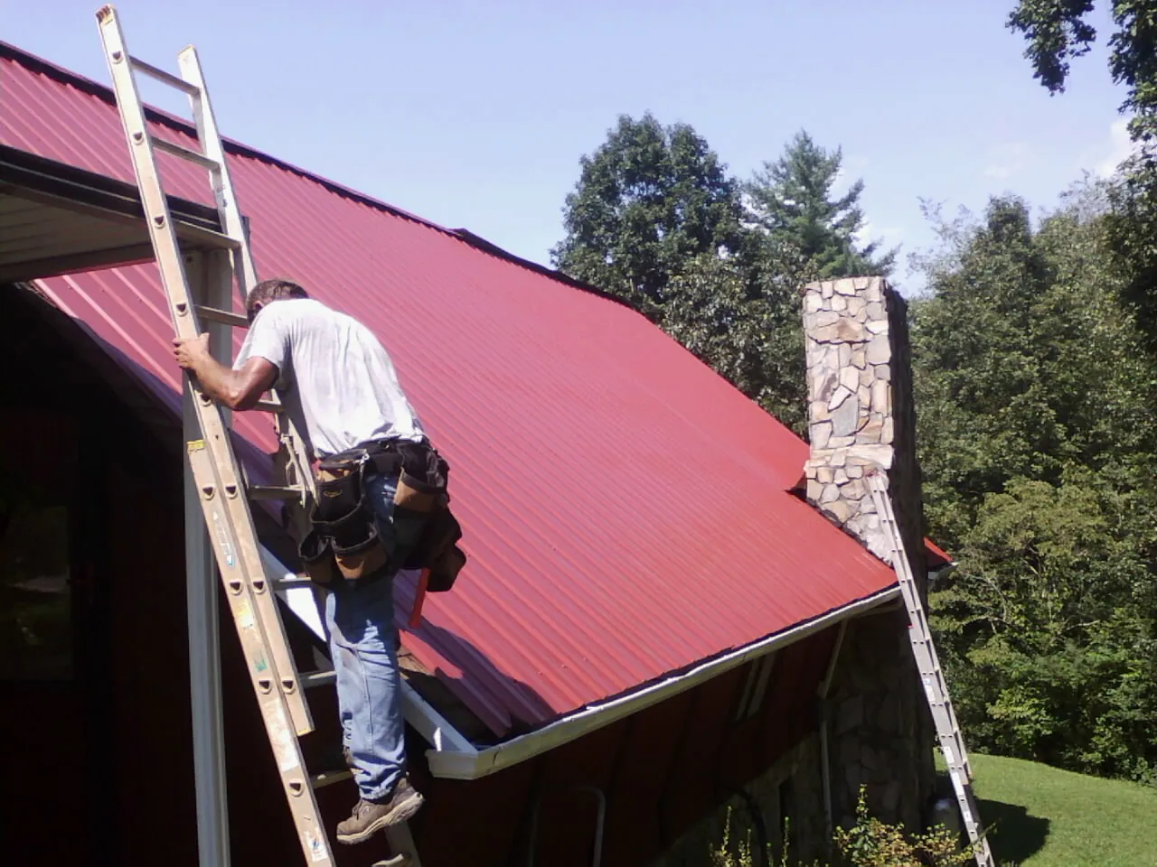 Roofer on a ladder beside a red metal roof and stone chimney — Soffit & Fascia Repair by Roofing Brothers in Firestone